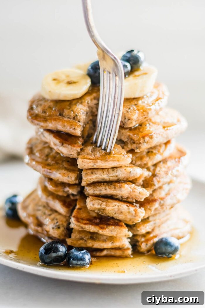 Fork digging into a big stack of chia seed pancakes on a plate. The pancakes are topped with banana, blueberries and maple syrup.
