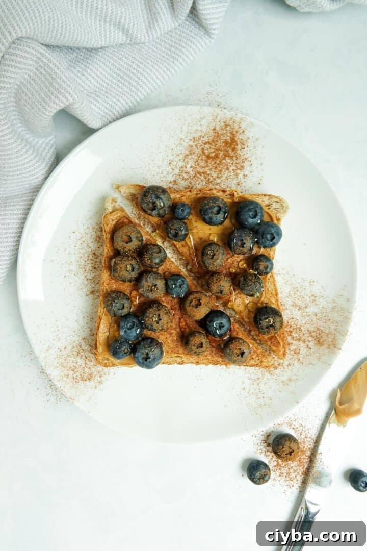 A slice of whole wheat toast with peanut butter, blueberries, cinnamon and honey displayed on a white plate on a white marble counter.