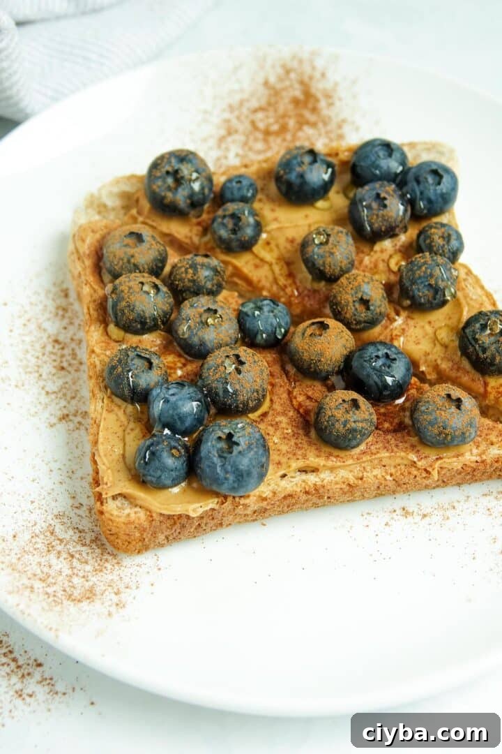 A close-up photos of slice of whole wheat toast cut in half, with peanut butter, blueberries, cinnamon and honey displayed on a white plate on a white marble counter.