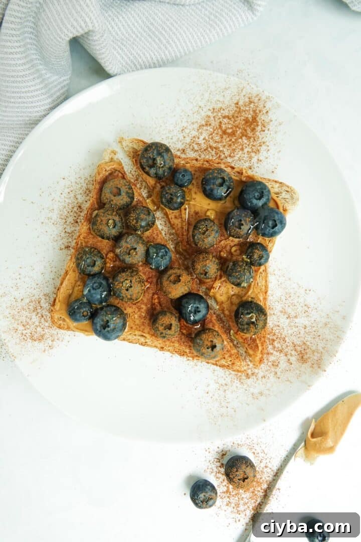 A slice of whole wheat toast with peanut butter, blueberries, cinnamon and honey displayed on a white plate on a white marble counter.