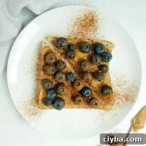 A slice of whole wheat toast with peanut butter, blueberries, cinnamon and honey displayed on a white plate on a white marble counter.