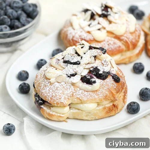A close-up of pastries filled with cream, topped with blueberries, almond slices, and powdered sugar on a white plate, with loose blueberries and a bowl of blueberries in the background.