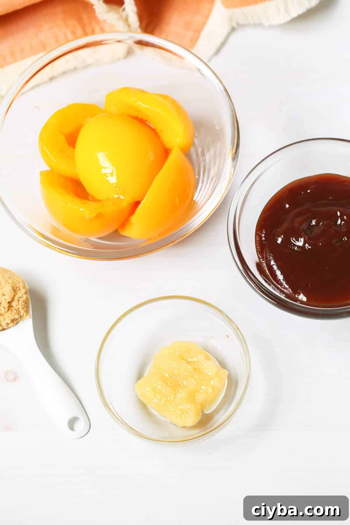 A clear bowl of peach slices, a clear bowl of Peach BBQ Sauce, a clear bowl of minced garlic, and a spoonful of brown sugar are arranged on a white surface.
