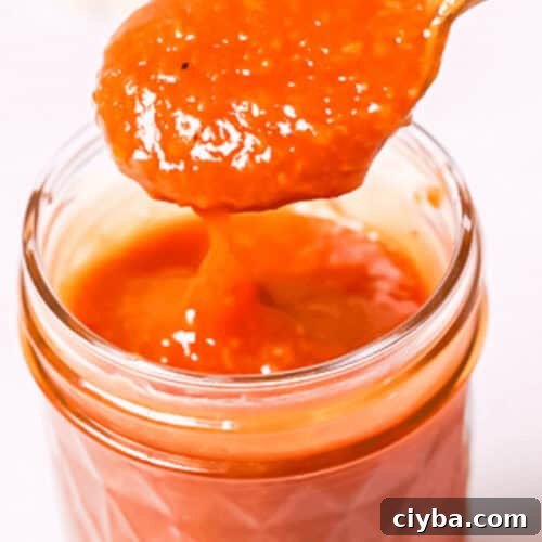 A spoon holding a portion of peach BBQ sauce is placed above a glass jar filled with the vibrant orange-red sauce.