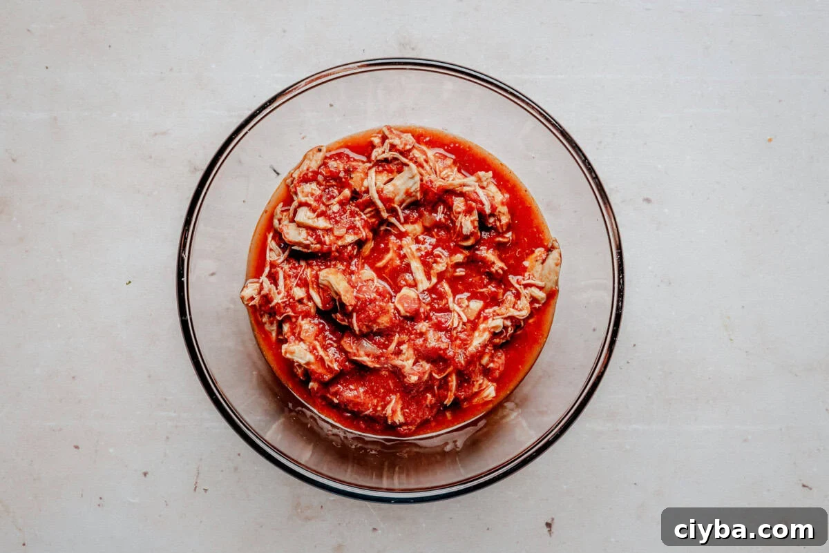 A clear glass bowl filled with shredded chicken in a red tomato-based sauce sits on a light-colored, textured surface.