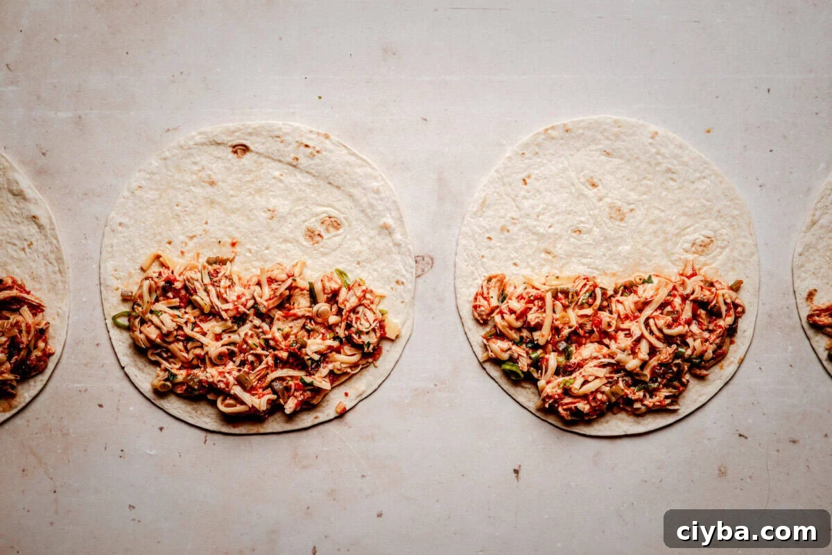 Four tortillas laid out on a light surface, each partially filled with a shredded chicken tinga, ready to be wrapped or rolled.