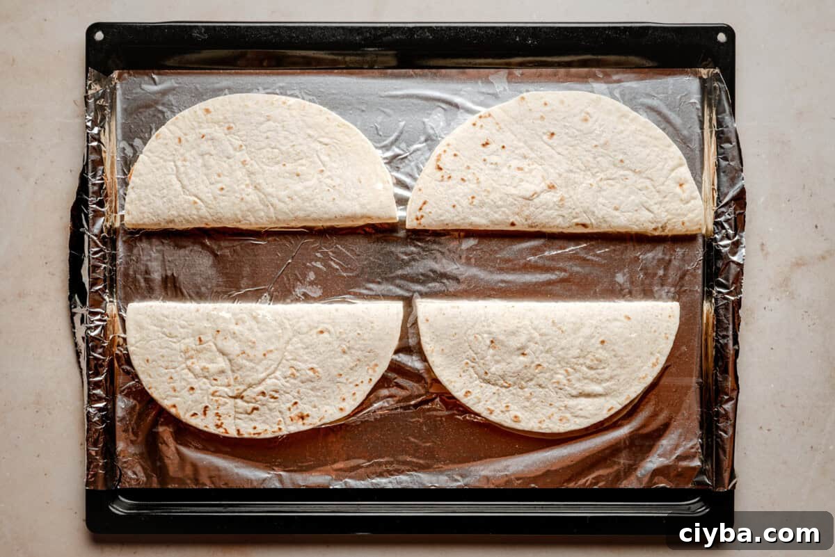 Four flour tortillas, each folded in half, are arranged on a baking sheet lined with aluminum foil. The baking sheet is placed on a light-colored countertop.