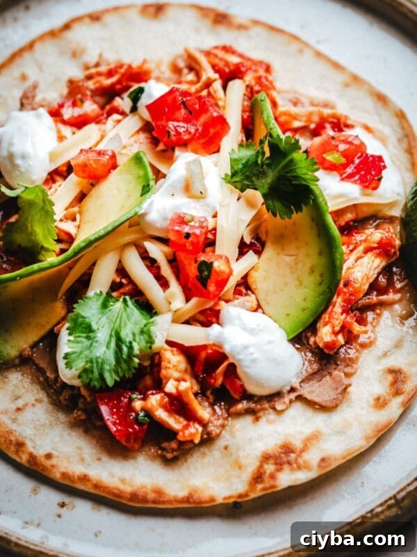 A close-up of a chicken tinga tostada topped with shredded chicken, sliced avocado, cheese, chopped tomatoes, fresh cilantro, and dollops of sour cream, served with a lime wedge on the side.