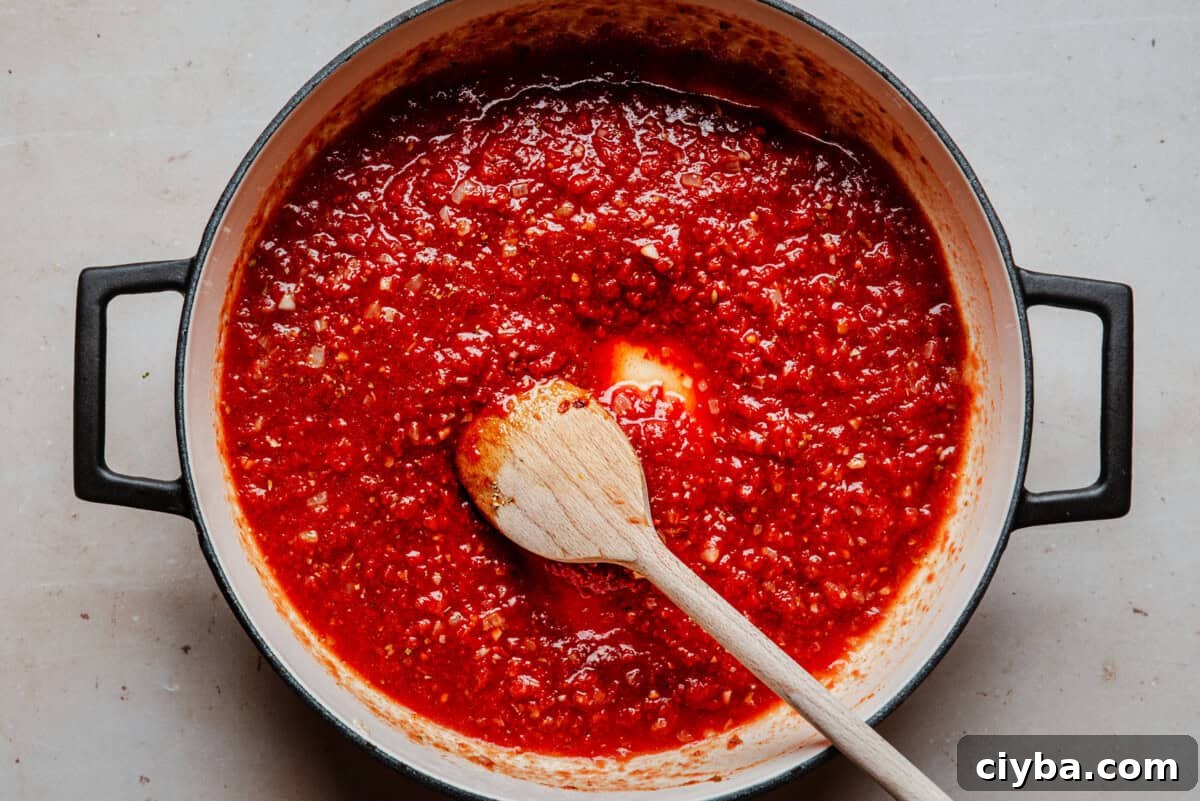 A pot of red tomato sauce simmering, with a wooden spoon resting inside, seen from above on a light-colored surface.