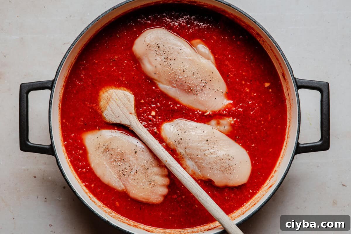 Raw chicken breasts sit in a pot of red tomato sauce, seasoned with pepper. A wooden spoon rests on the side, and the pot has black handles. The scene is viewed from above on a light surface.