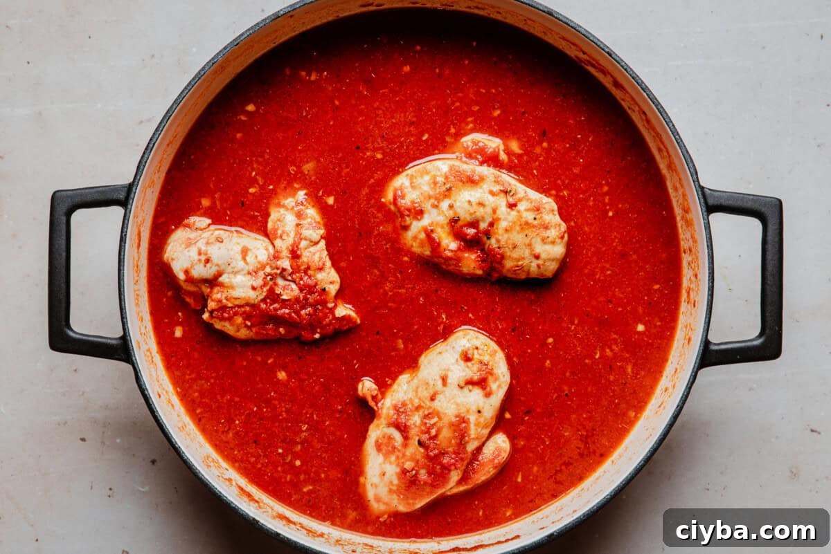 A pot with three pieces of chicken simmering in a thick, red tomato-based sauce, viewed from above on a light-colored surface.