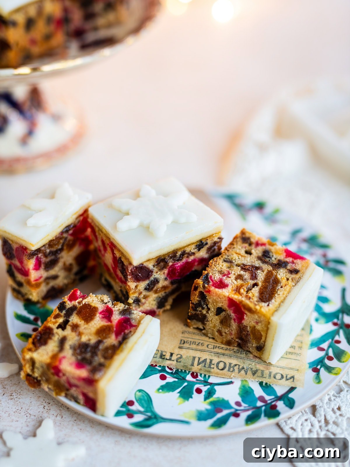 Four pieces of fruitcake topped with white icing and snowflake decorations are arranged on a festive plate. The fruitcake is filled with colorful candied fruit and nuts. A cake stand and more fruitcake are in the background.