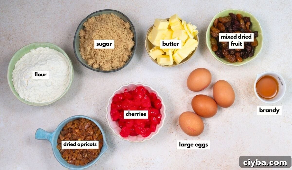 Ingredients for baking Mary Berry Christmas cake bites are displayed in bowls on a white surface: flour, brown sugar, butter, mixed dried fruit, brandy, large eggs, dried apricots, and glacé cherries—each bowl neatly labeled.