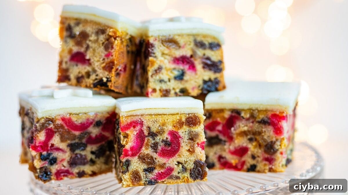 Seven square slices of fruitcake with white icing are displayed on a glass cake stand, with two more slices on plates nearby. The cake is filled with colorful dried fruit. Soft, warm lights blur in the background.