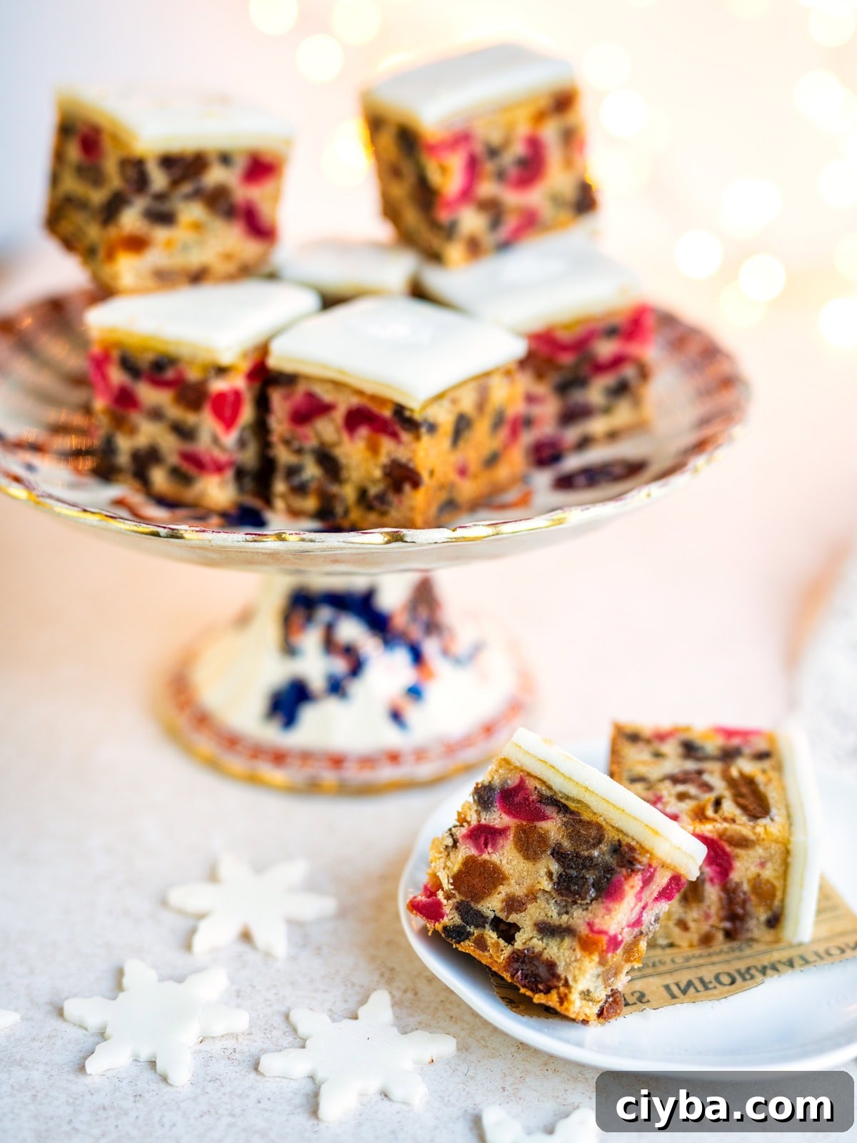 Small squares of fruitcake with white icing are displayed on a decorative cake stand and a plate. Edible snowflake decorations are on the table, with bright, festive lighting in the background.