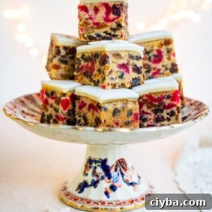 A decorative cake stand holds a stacked arrangement of Christmas Cake Bites topped with white icing, displaying colorful pieces of candied fruit and nuts. Soft lights twinkle in the blurred background.