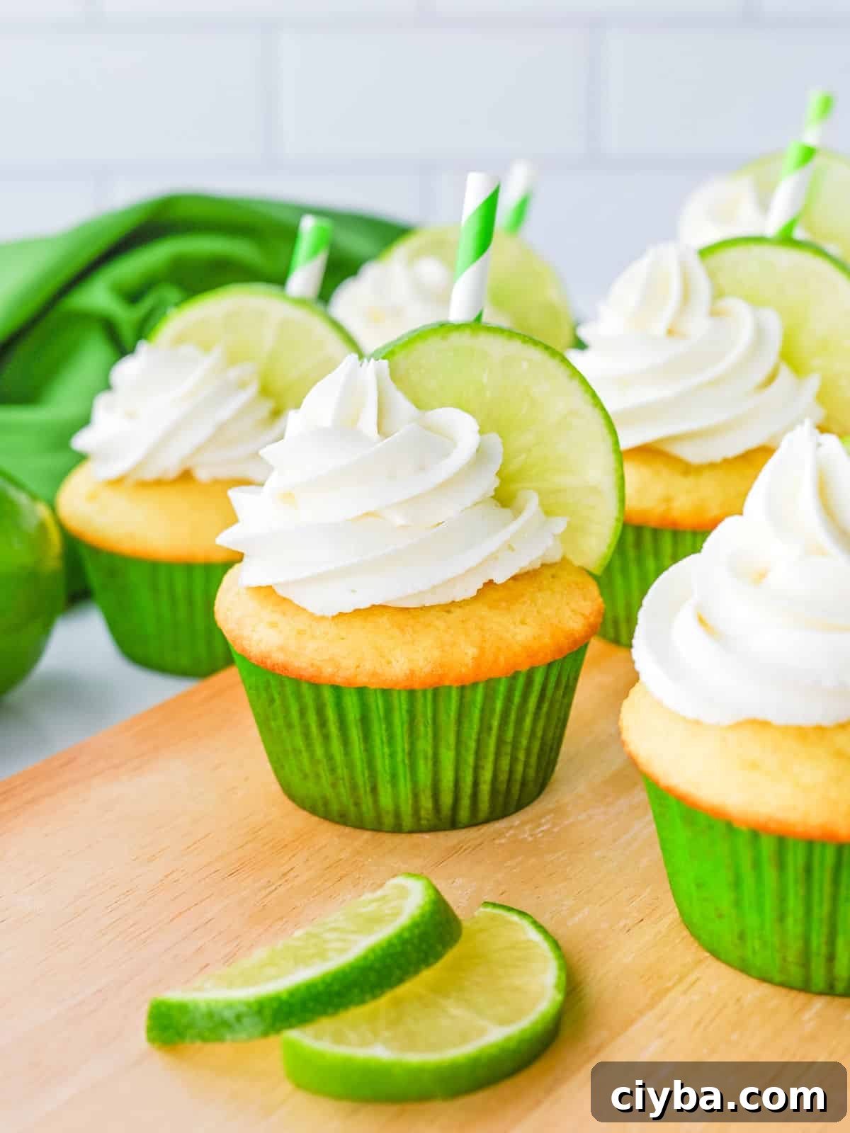Cupcakes with white frosting, lime slices, and green-striped straws are arranged on a wooden surface, with extra lime slices in front and a green cloth in the background.