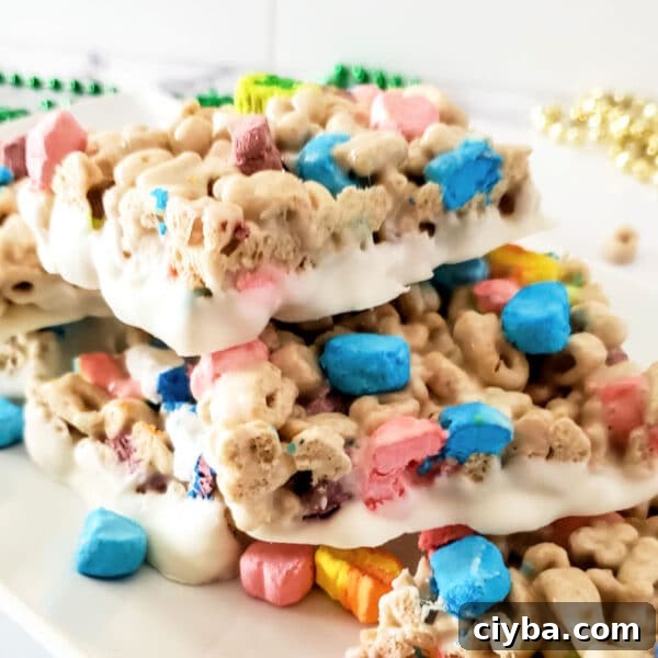 Three stacked cereal bars with colorful marshmallows and a white coating are displayed on a white plate, with green and gold bead necklaces in the background.