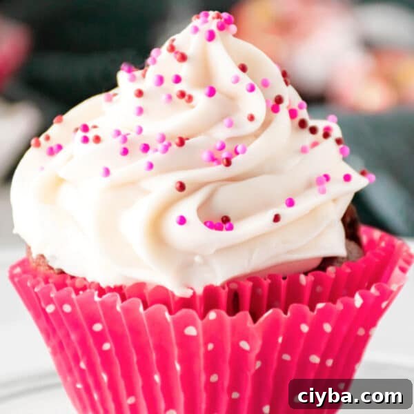 A cupcake in a pink polka dot wrapper topped with white frosting and pink and red sprinkles.