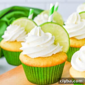 Vanilla cupcakes with white frosting, garnished with a lime slice and green-striped straw, in green paper liners on a wooden surface.