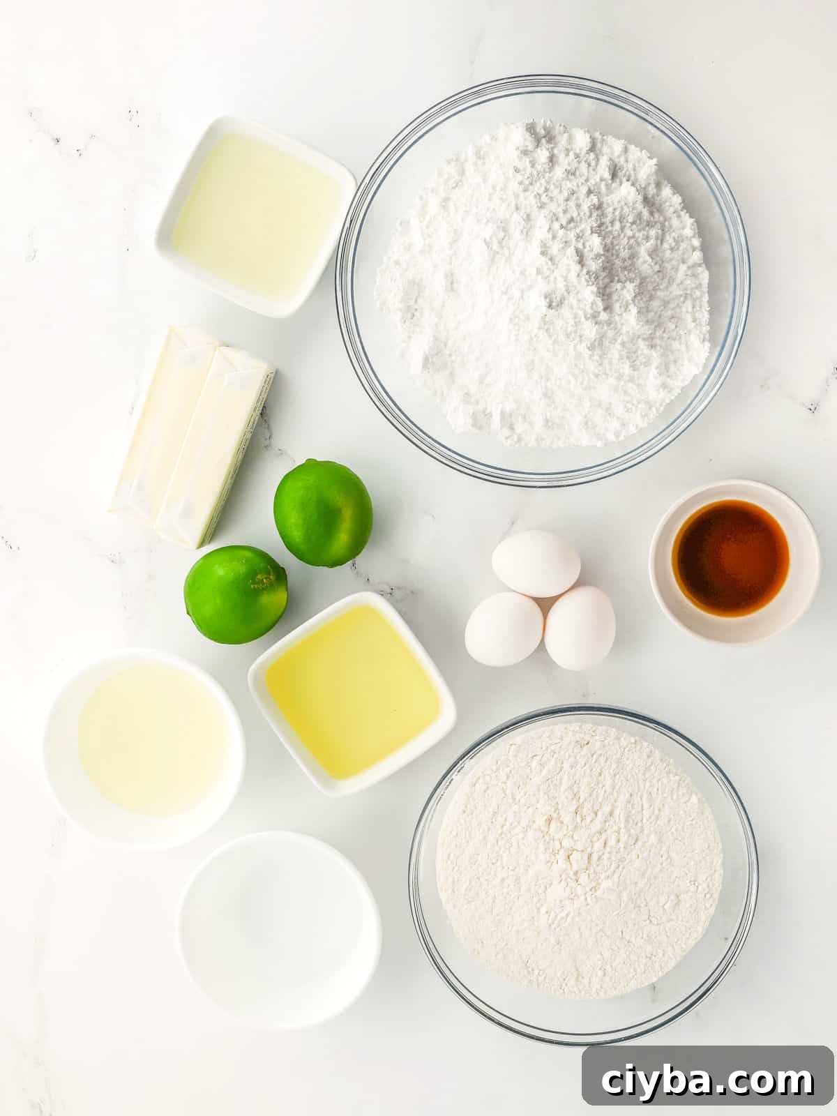 Various baking ingredients including flour, powdered sugar, eggs, limes, butter, oil, vanilla extract, and other liquids arranged in bowls on a white surface.