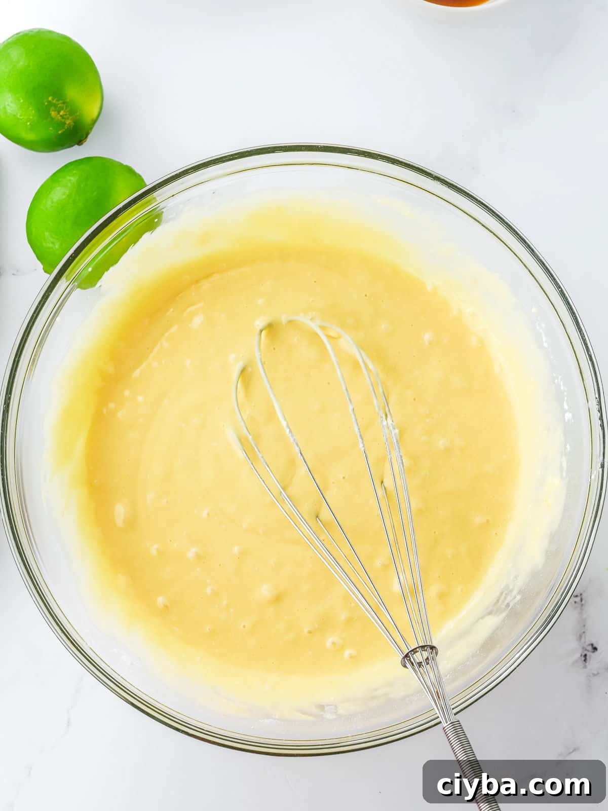 A glass bowl of yellow cake batter with a metal whisk inside, placed on a white surface next to two whole limes.
