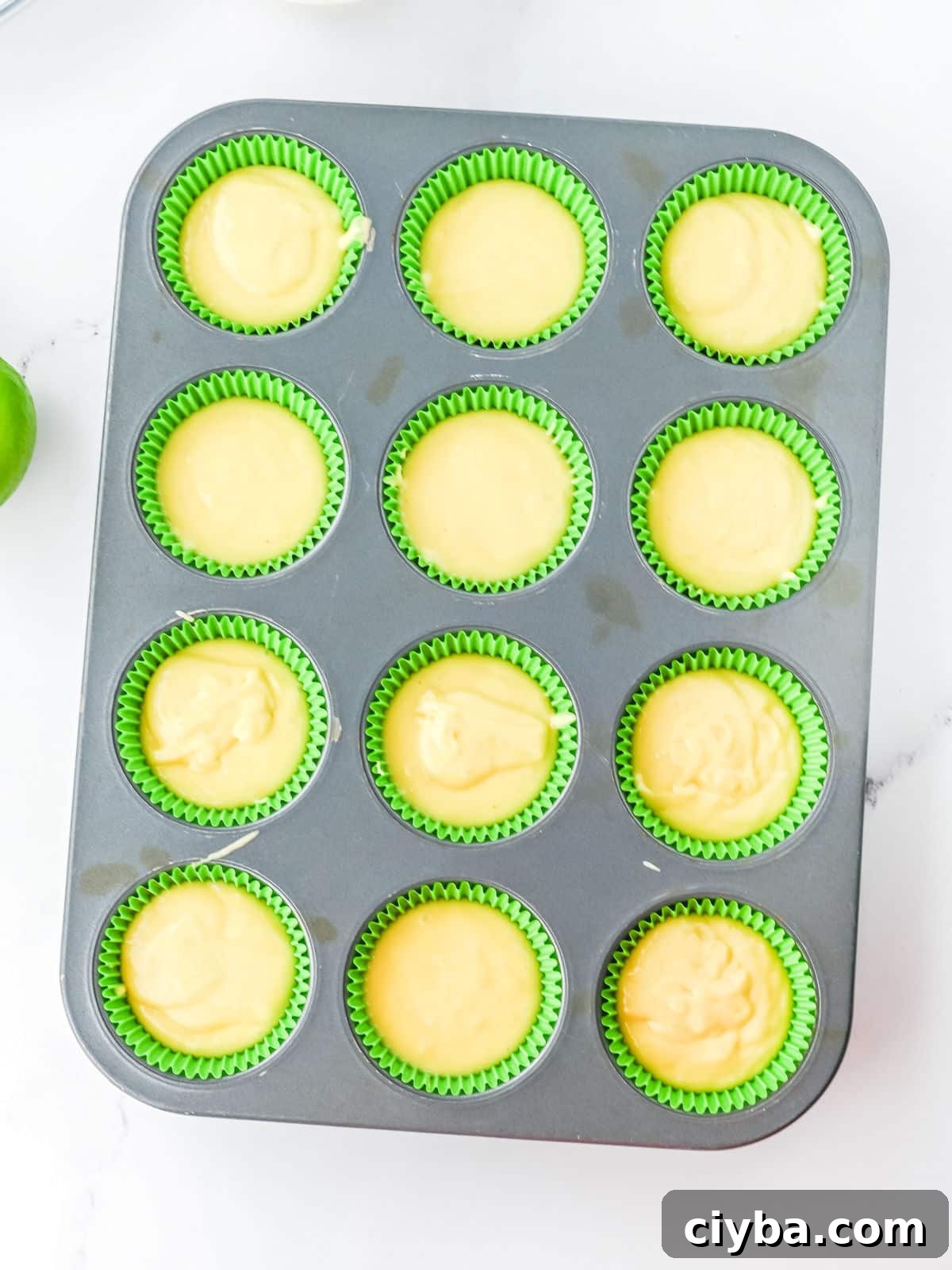 A muffin tin lined with green paper cups, each filled with yellow batter, sits on a white surface.