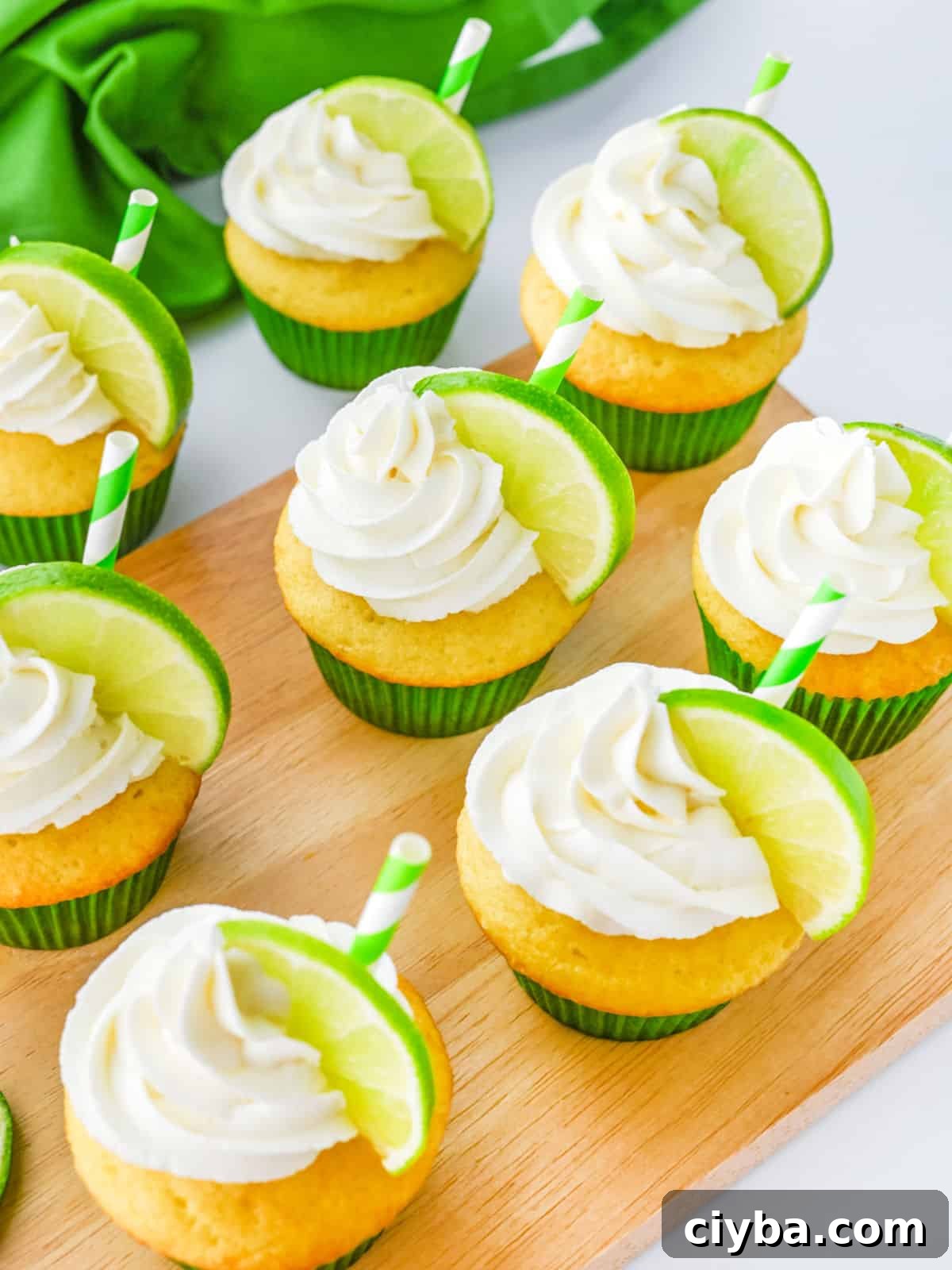 Cupcakes with white frosting, each topped with a lime wedge and a green-striped straw, arranged on a wooden board with a green cloth in the background.