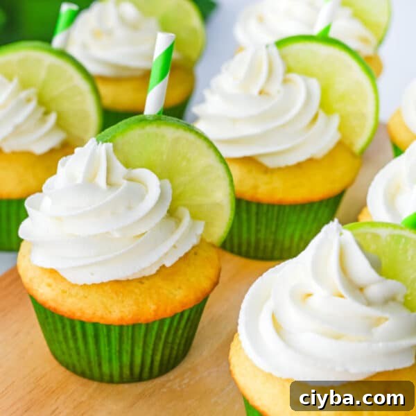 Several cupcakes with white frosting, each topped with a slice of lime and a green-striped paper straw, sit on a wooden surface.