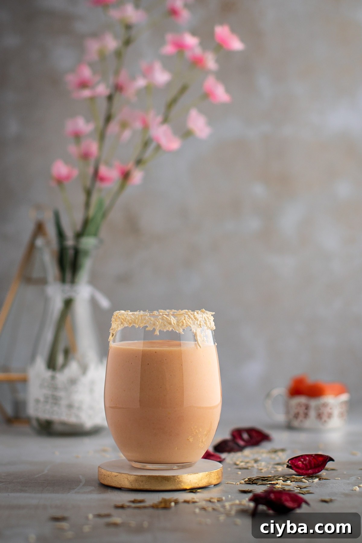 A glass of light pink papaya smoothie on a gold coaster, with pink flowers in the background.