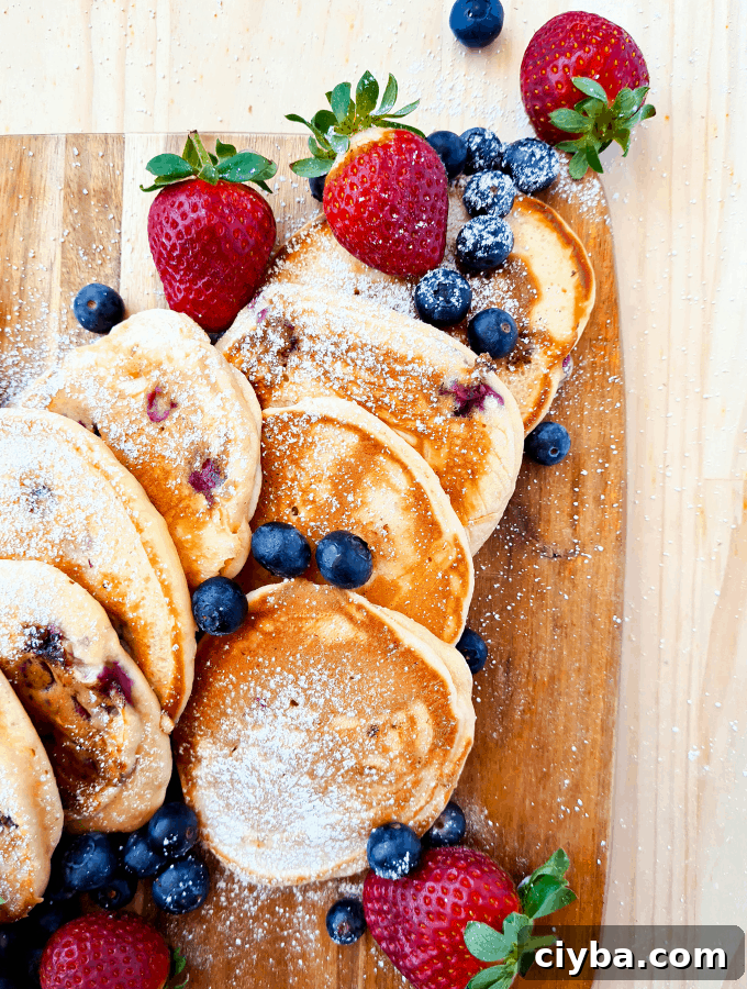 Pancakes sprinked with icing sugar/powder on top of a wooden board, decorated with blueberries and strawberries
