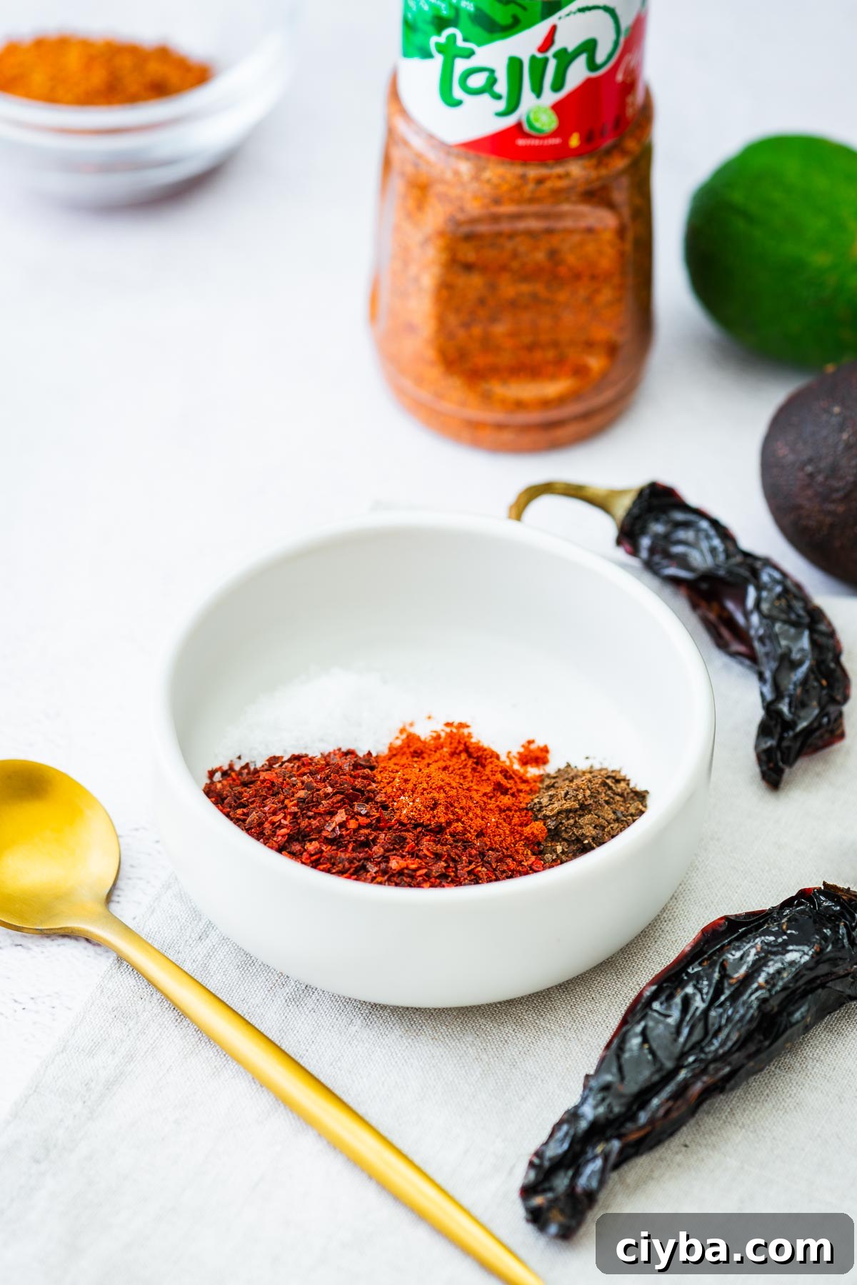 The ingredients for a tajin substitute blend in a small white bowl, including smoked paprika, chilli flakes, black lime powder, salt and citric acid.