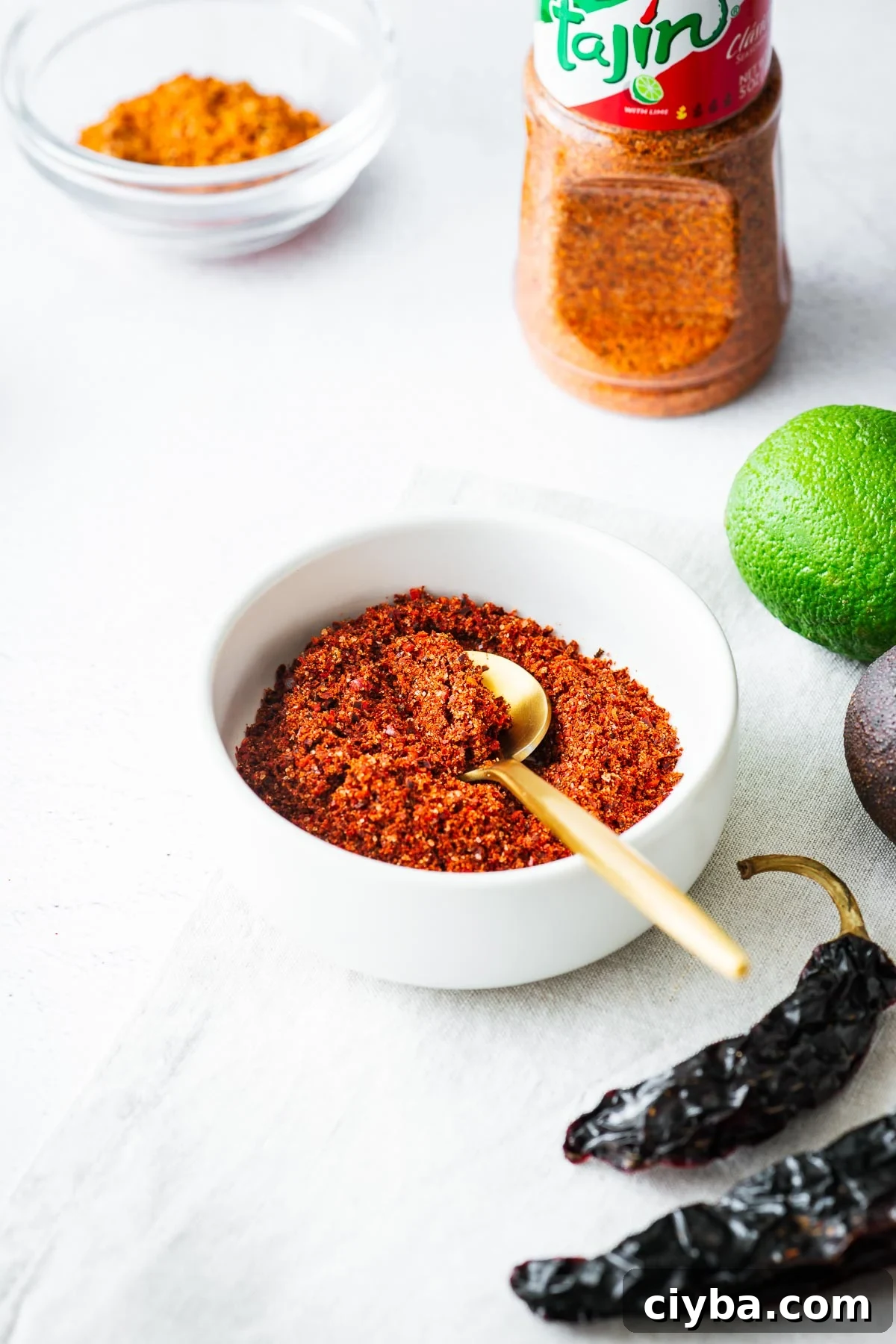 A homemade spice blend of chilli flakes, citric acid, dehydrated limes and salt in a small white bowl with a bottle of Tajín Clásico Seasoning in the background.