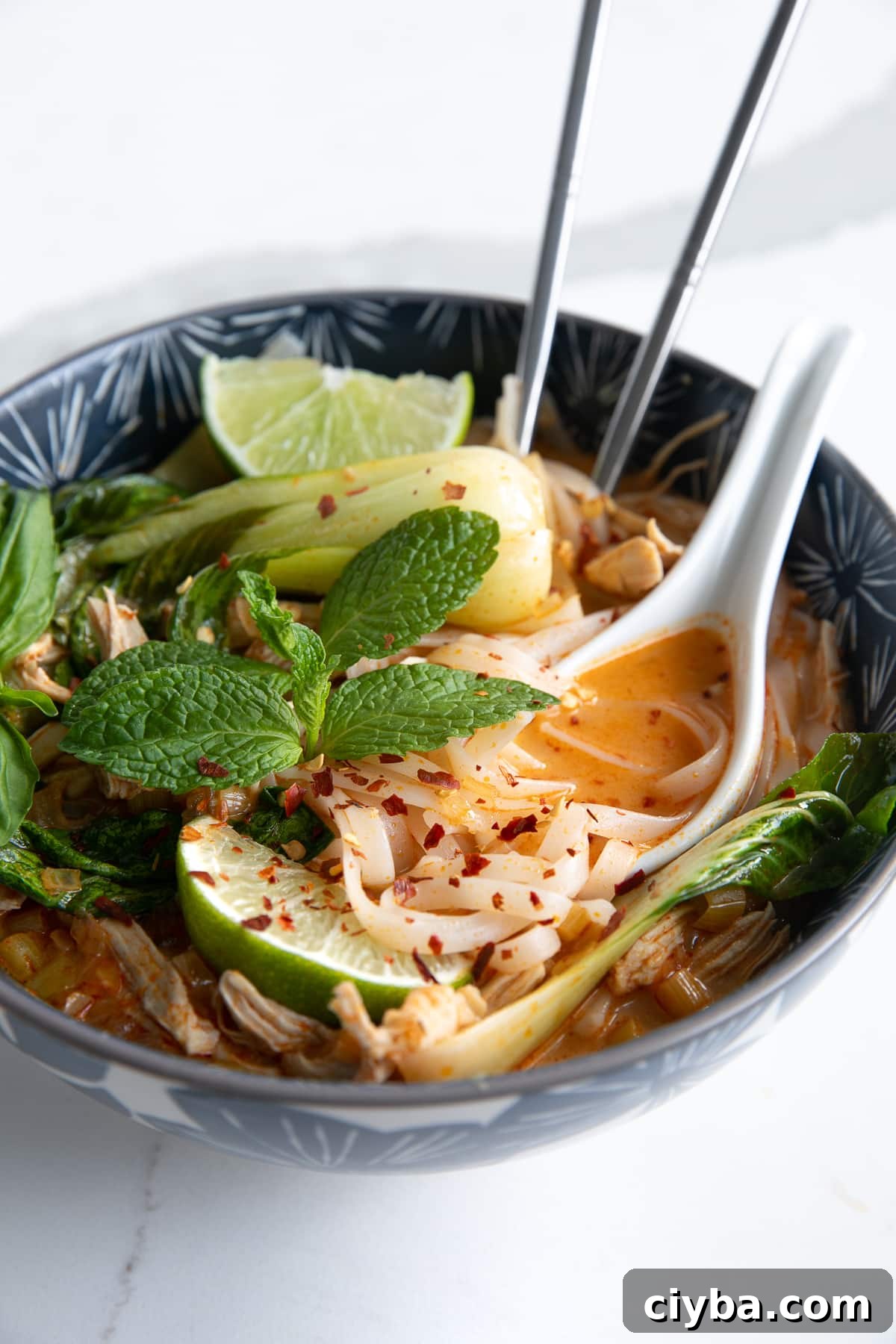 Thai Chicken Curry Noodle Soup: Authentic Recipe & Tips 6 Image of a blue soup bowl filled with Thai chicken curry noodle soup made with a coconut curry broth, rice noodles, shredded chicken, and bok choy, and garnished with fresh mint, limes, and fish sauce.
