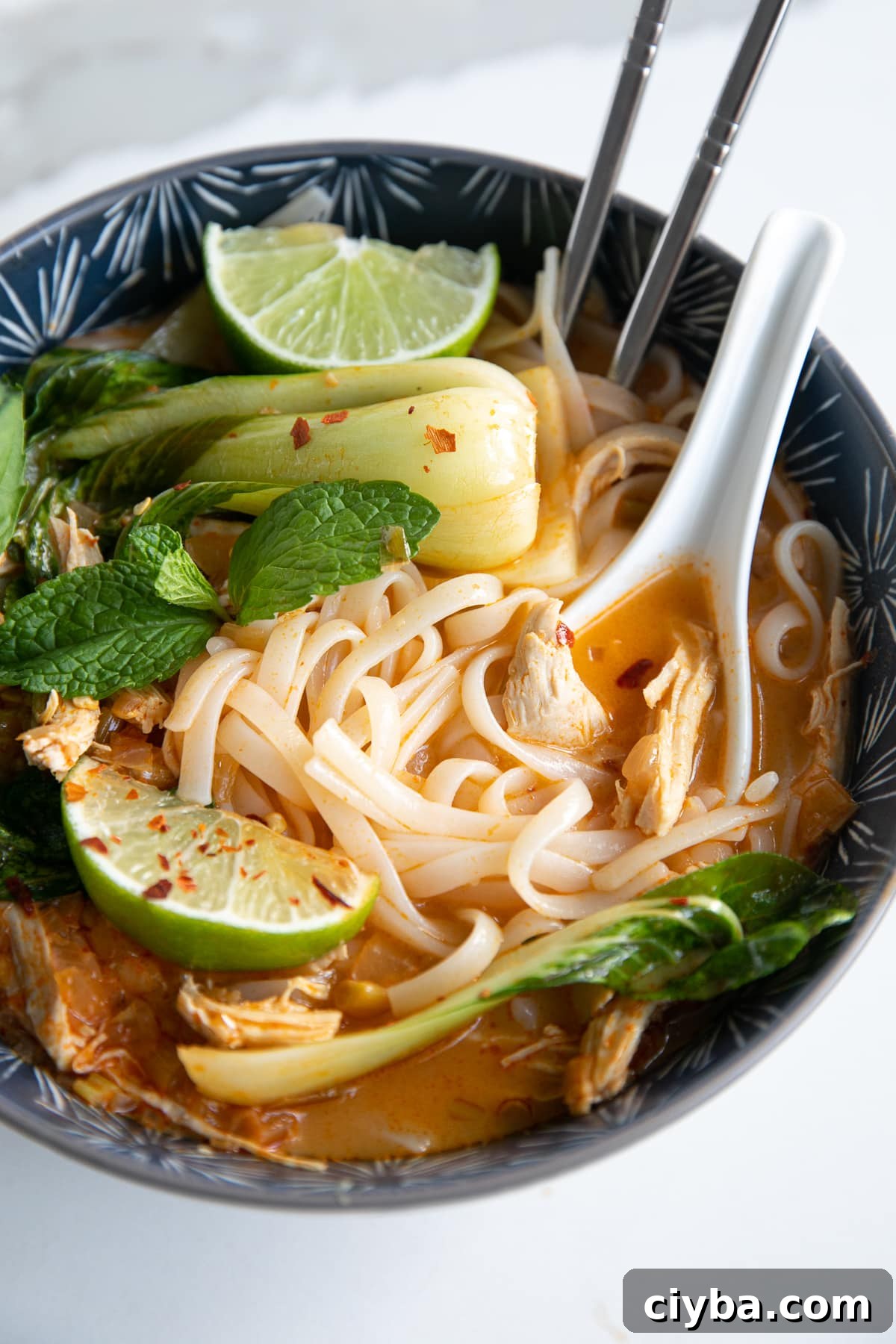 Thai Chicken Curry Noodle Soup: Authentic Recipe & Tips 7 Image of a blue soup bowl filled with Thai chicken curry noodle soup made with a coconut curry broth, rice noodles, shredded chicken, and bok choy, and garnished with fresh mint, limes, and fish sauce.