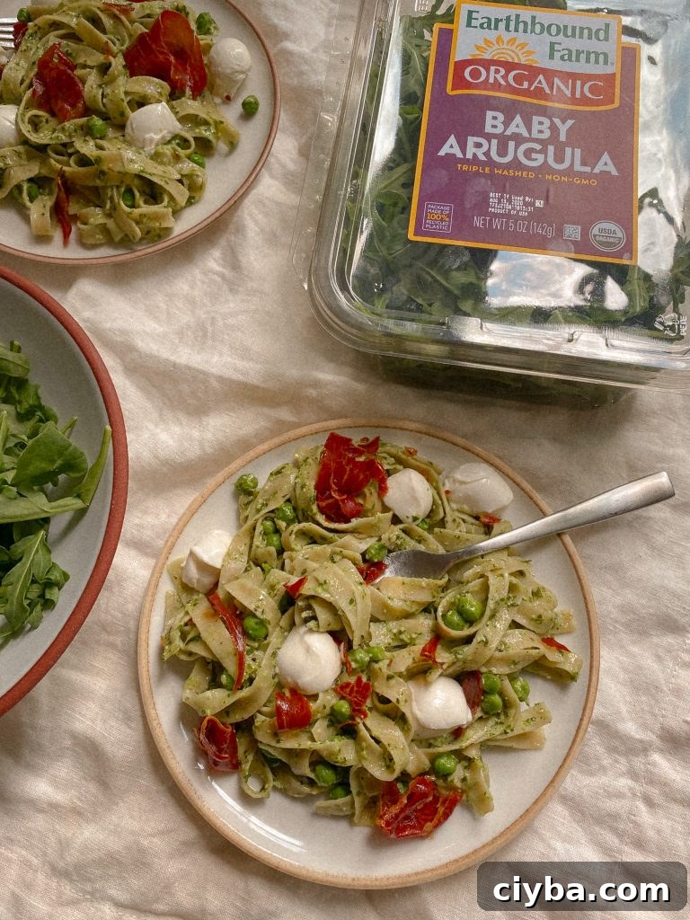 pesto pasta with prosciuttio, mozzarella, and peas on a plate with fork and bowl of arugula 