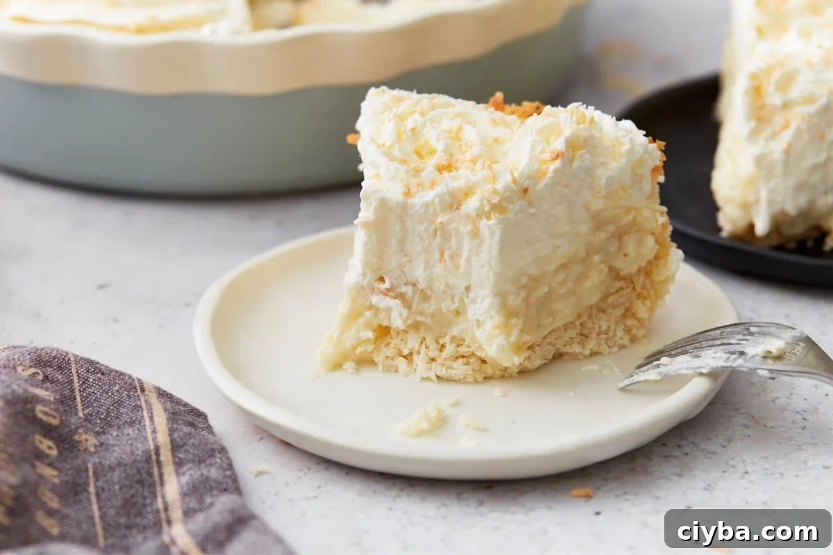 Close-up of a single slice of coconut cream pie on a white plate, with toasted coconut on top and a fork nearby.