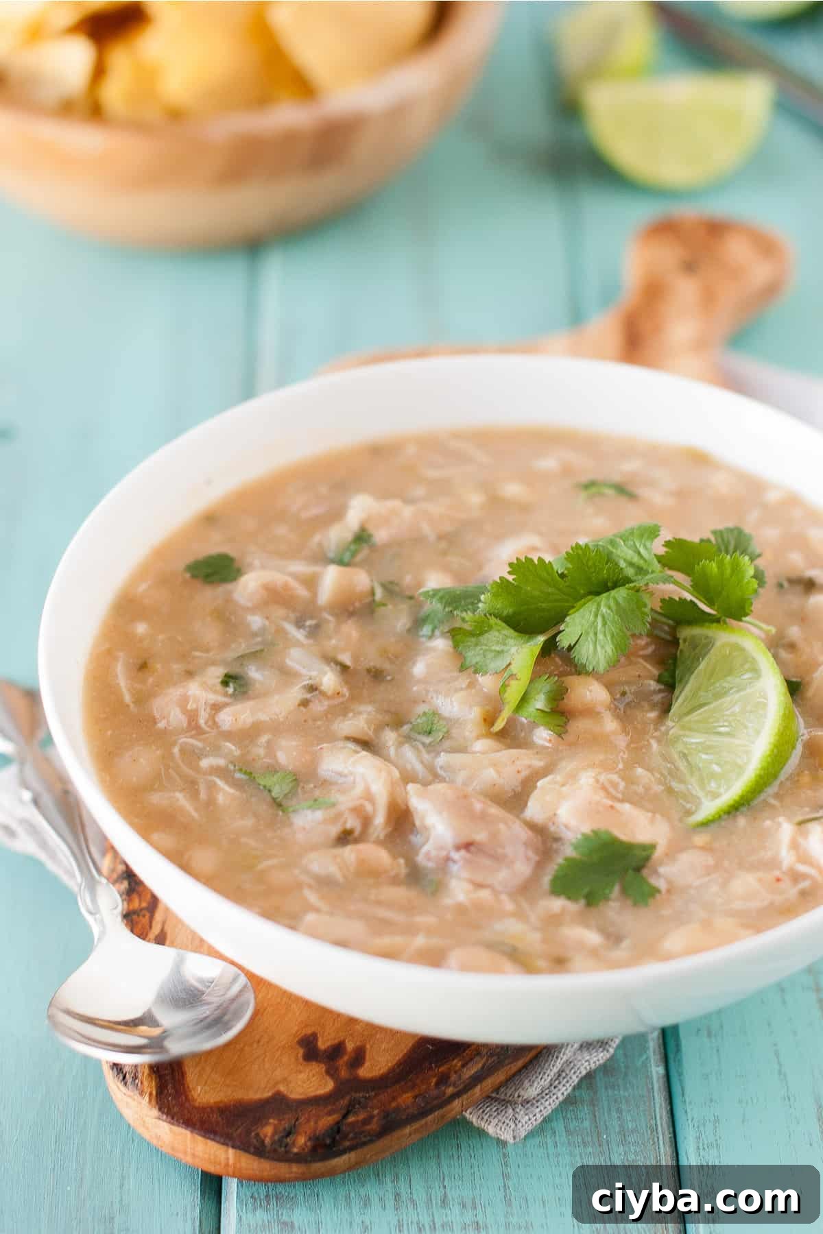 Healthy white chicken chili in a white bowl on a green background on a brown cutting board with a spoon, topped with lime and cilantro.