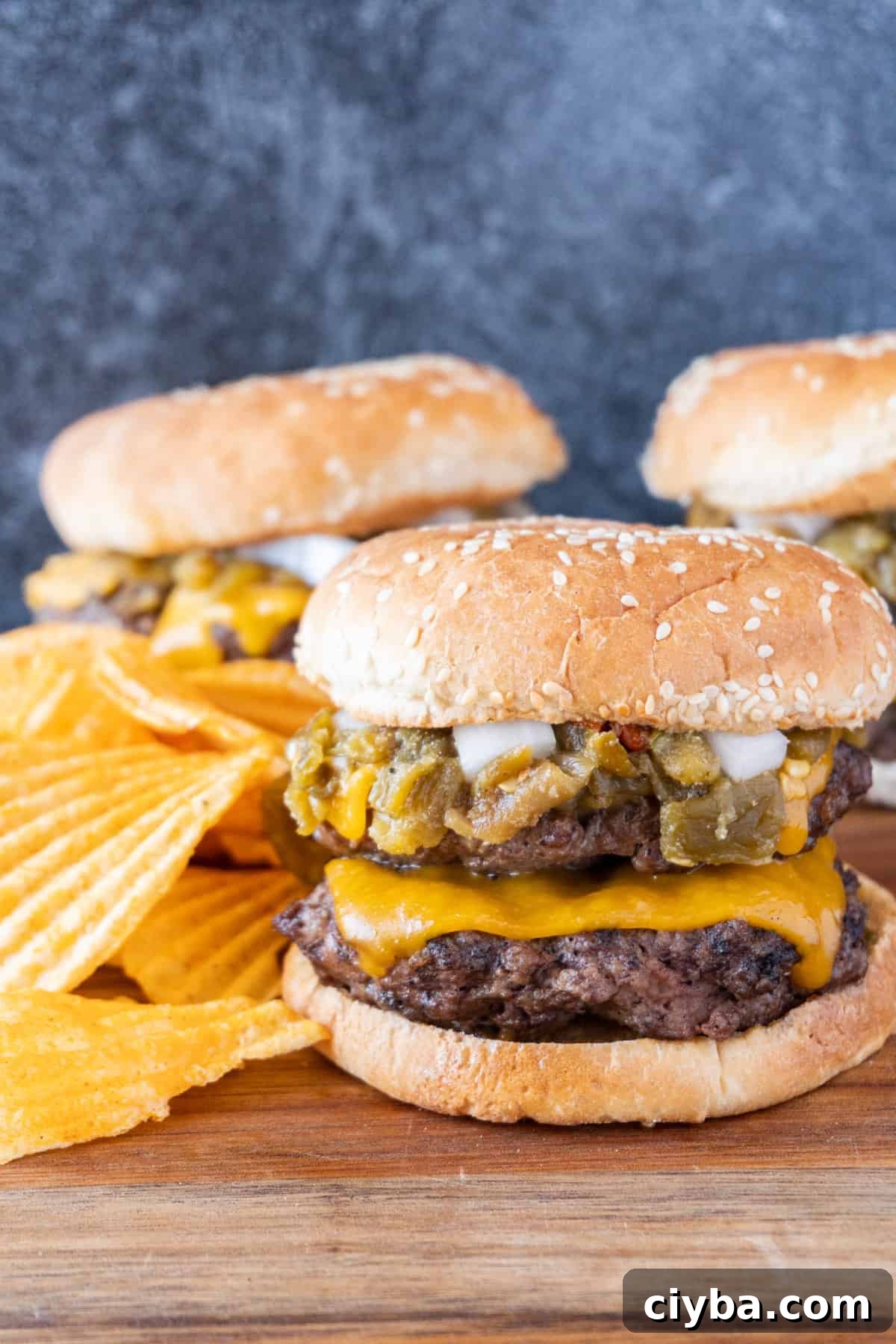 Green Chile Cheeseburger, 2 patties with American cheese, green chile sauce, and onions, next to chips on a wooden cutting board.