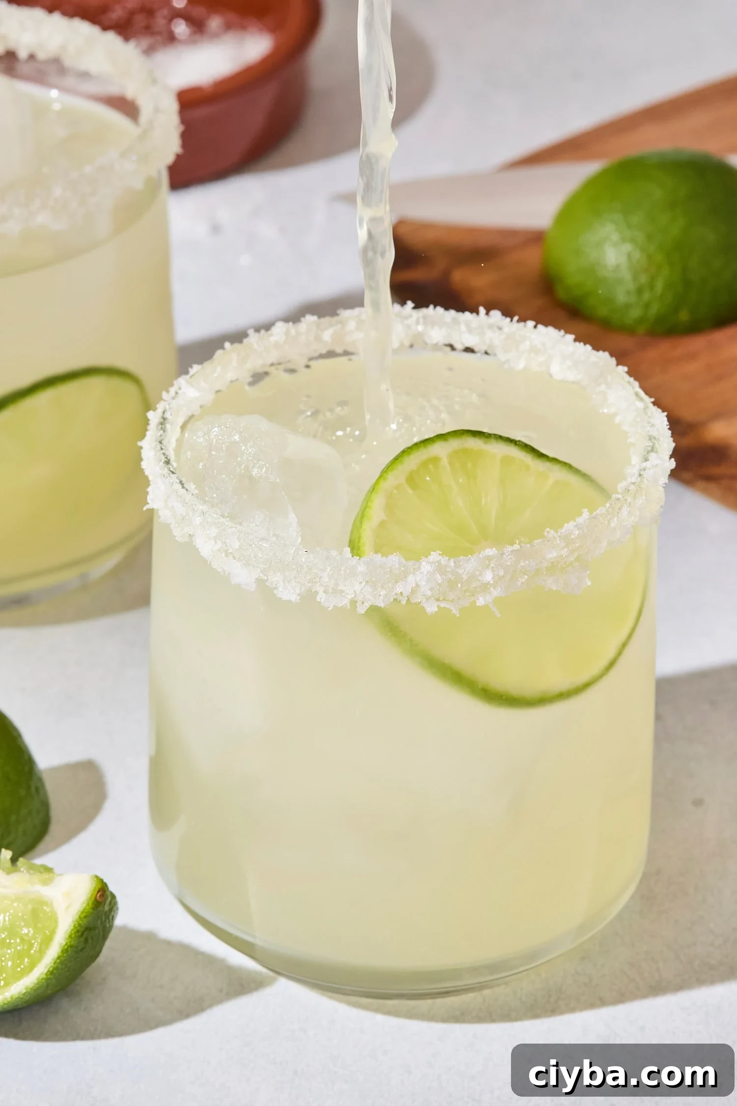 Classic Margarita Recipe: How to Make an Authentic Margarita 3 Margarita being poured into a glass with light green margarita already in it with a lime slice.