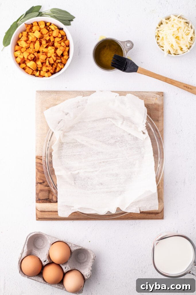 A pie dish lined with the first sheet of phyllo dough on a wooden board surrounded by bowls of squash, shredded cheese, sage leaves, eggs, olive oil, and milk.