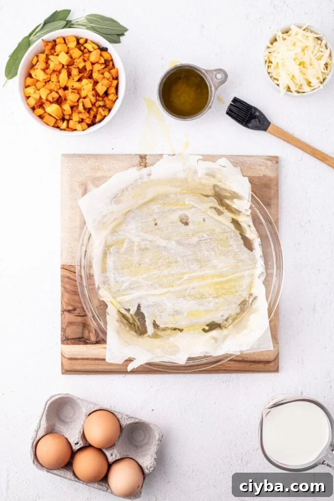 Close up of phyllo dough brushed with olive oil in a pie dish, with ingredients like squash, cheese, eggs, milk, and sage nearby.
