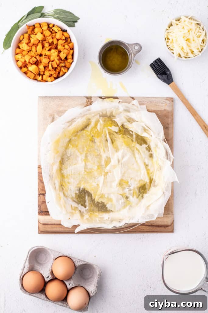 Multiple layers of phyllo dough brushed with olive oil in a pie dish, surrounded by roasted squash, shredded cheese, eggs, and milk.
