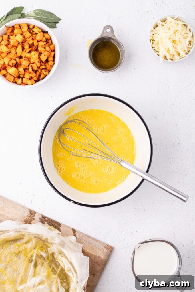 Mixing bowl with beaten eggs and whisk, with nearby bowls of roasted squash, shredded cheese, olive oil, and a prepared phyllo crust.