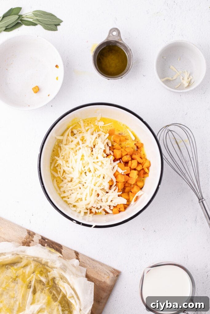 Mixing bowl filled with beaten eggs, roasted squash, and shredded Gruyère cheese, with whisk and prepared crust nearby.
