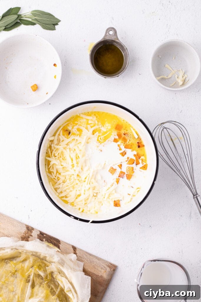 Egg mixture in bowl with added half and half, roasted squash, and shredded Gruyère cheese, with whisk and phyllo crust nearby.