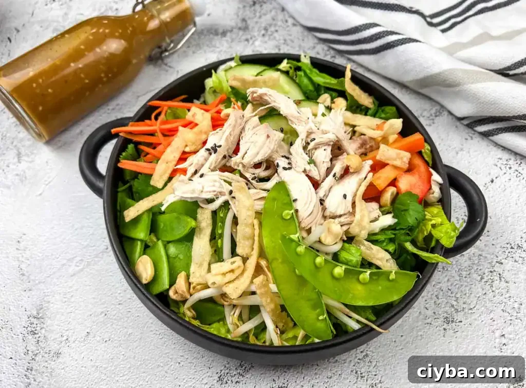 A bowl of Chinese chicken salad with shredded chicken, snap peas, carrots, and peppers, accompanied by a bottle of dressing on the side.