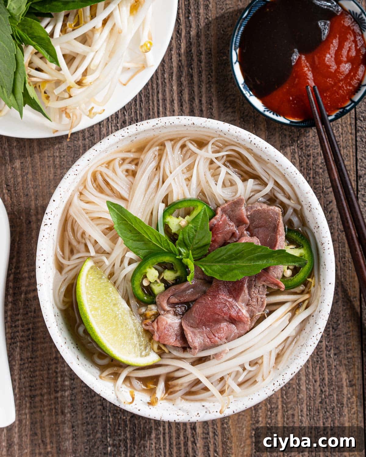 Bowl of pho tai with lime wedges, herbs, hoisin sauce, and sriracha.