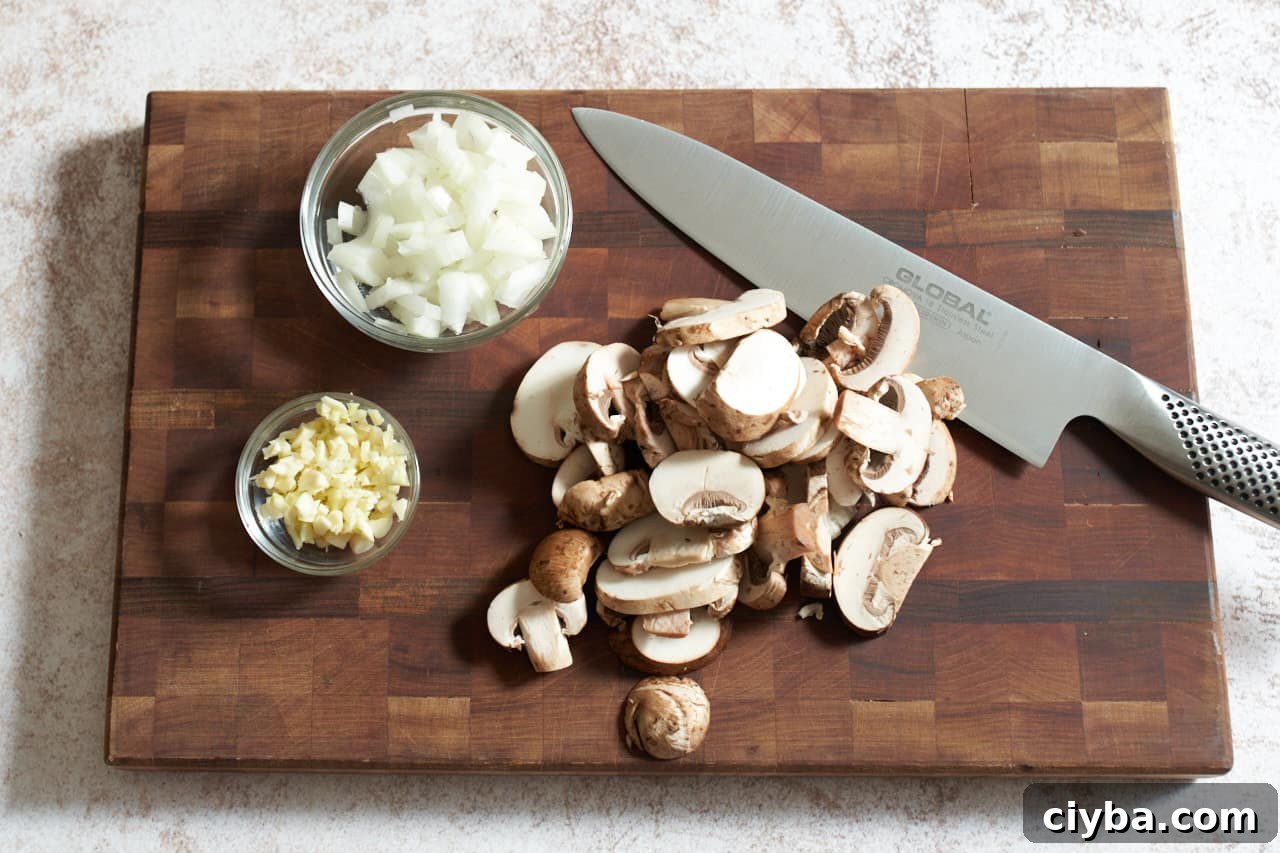 A knife on a cutting board with sliced mushrooms and two small bowls of diced onions and garlic.