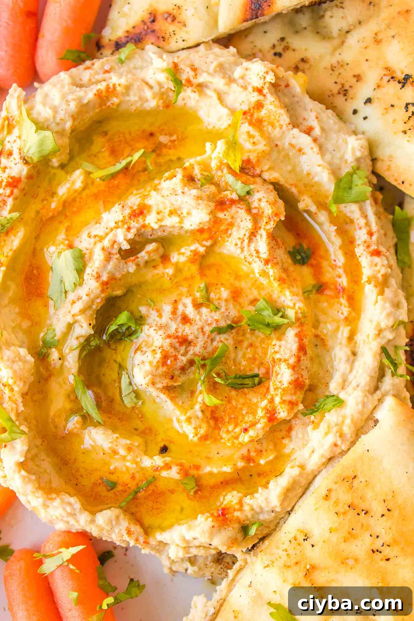 Creamy hummus drizzled with olive oil surrounded by wedges of pita and baby carrot.